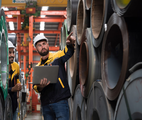 male factory worker using laptop computer inspecting quality rolls of galvanized or metal sheet rolls